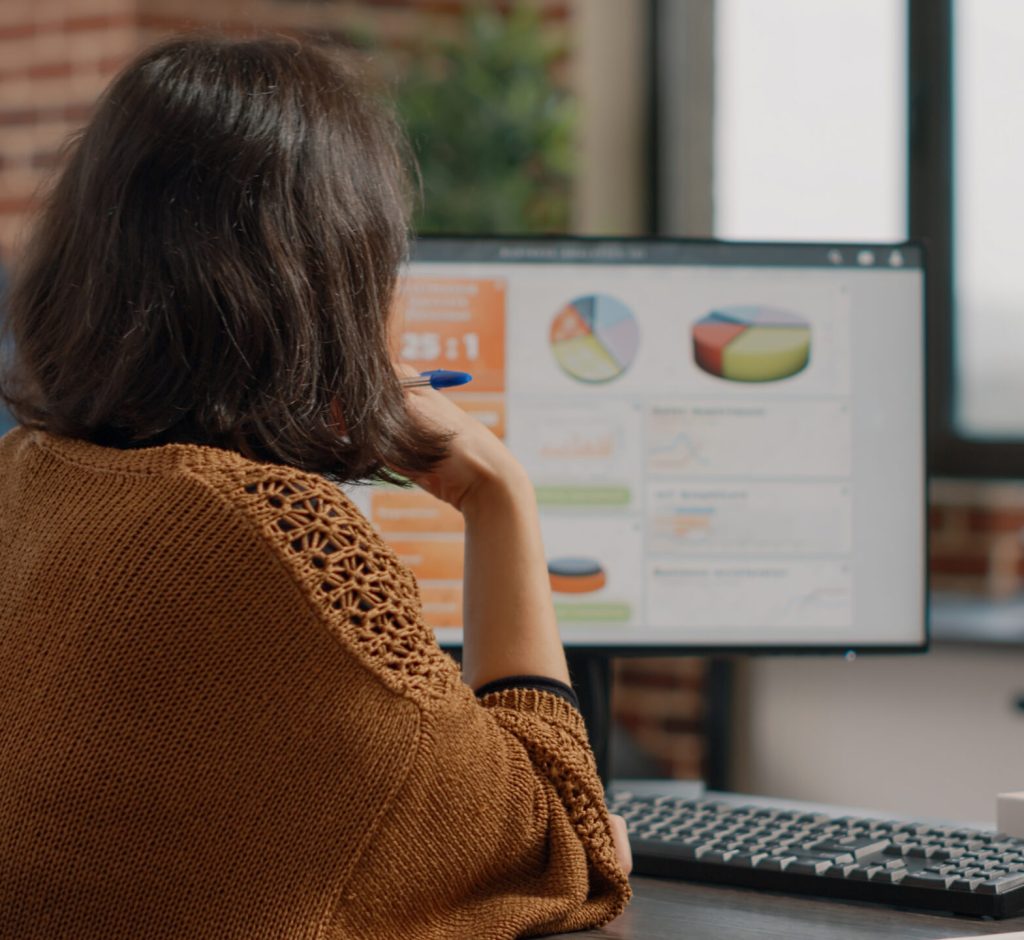 Business woman analyzing rate charts on computer and files to plan project and strategy. Employee using information on monitor and documents to design marketing presentation at desk.